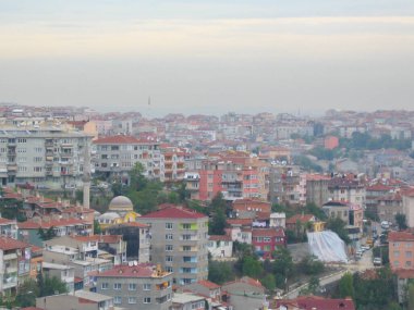 Sabahın erken saatlerinde canlı bir şehrin panoramik bir görüntüsü, kırmızı çatıları, yeşil alanları ve yumuşak bir gökyüzünün altındaki uzak tepelerin ipuçlarını gösteren renkli binalar..