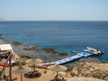 Güzel bir kıyı sahnesinde açık mavi denize uzanan ahşap bir rıhtım vardır. Şezlonglar kumlu kayalara oturur, sıcak bir yaz gününde güneşin altında dinlenmeye davet ederler..