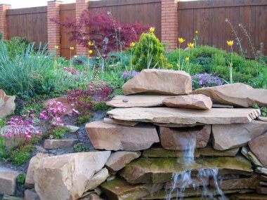 Huzurlu bir bahçe, canlı çiçekler ve gür yeşillikler arasında yavaşça çağlayan büyüleyici bir taş su özelliğini sergiler. Ahşap çit mahremiyet ve dinlendirici dış uzaya kırsal bir dokunuş ekler..