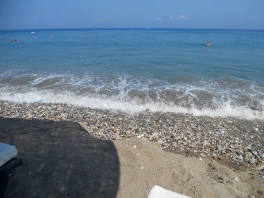 Dalgalar, insanların güneşli kumsalın keyfini çıkardıkları çakıl taşlarına hafifçe çarpar. Açık mavi su ufka kadar uzanıyor ve dinlenmek için mükemmel sakin bir atmosfer yaratıyor..