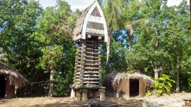 Uzun, karmaşık bir şekilde tasarlanmış totem yoğun ağaçların ve güneş ışığı altında geleneksel kulübelerin arasında duruyor..