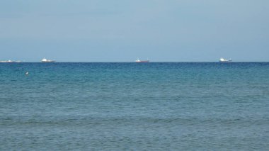 Huzurlu bir okyanus manzarası yumuşak dalgalar ve açık mavi bir gökyüzü içerir. Uzaktaki kargo gemileri ufukta görünüyor ve sakin deniz manzarasına ilgi gösteriyor..