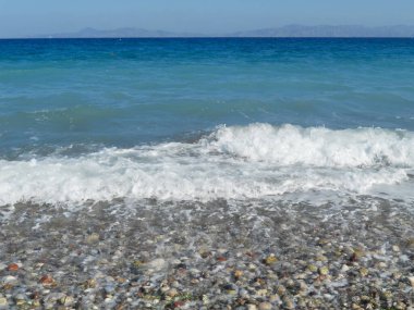 Yumuşak dalgalar, berrak mavi gökyüzünün altında kıyıdaki renkli çakıl taşlarının üzerinden süzülerek sakin bir sahil atmosferi yaratır. Güneş ışıl ışıl parlıyor, rahatlatıcı ve keyifli..