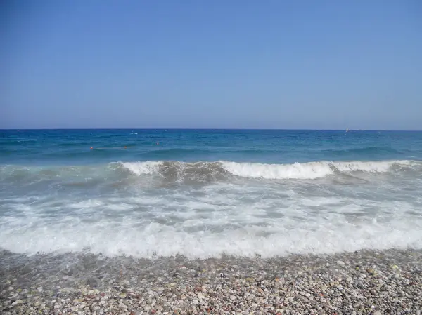 Muhteşem dalgalar, berrak mavi gökyüzünün altındaki çakıllı bir plaja çarpıyor. Deniz, mavi ve yeşilin çeşitli tonlarını yansıtırken güneş ışıl ışıl parlıyor ve sakin bir kıyı atmosferi yaratıyor..