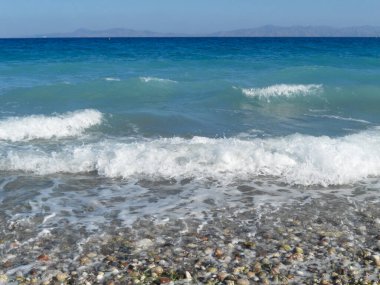 Renkli çakıl taşlı plajda hafif dalgalar çarpar. Açık gökyüzü bu sakin kıyı manzarasına zemin hazırlıyor..