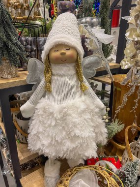 Tüylü beyaz bir elbise ve rahat bir şapkayla süslenmiş büyüleyici bir peri bebeği duruyor. Yumuşak detaylar ve meleksi özellikler samimi bir tatil gösterisinde neşeli bir atmosfer yaratıyor..