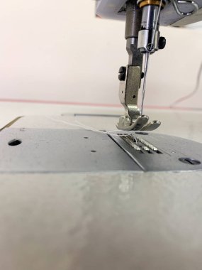 Dikiş makinesi etkin bir şekilde kumaş dikiyor, iğneyi ve ayağı keskin detaylarla gösteriyor. Dekor bir atölye, işçiliğin ve dikiş dikmeye adanmışlığın vurgulandığı.