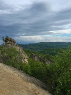 Dağ tepesinde, büyük bir kaya oluşumu ön plana hakim olur. Aşağıdaki manzarada yemyeşil ağaçlar, bulutlarla dolu dramatik bir gökyüzünün altında, değişen havayı ima ederek uzanıyor..