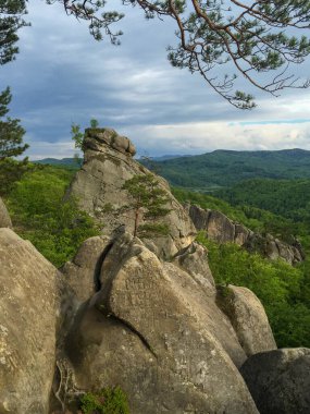 Kayalık bir çıkıntı, yemyeşil ağaçlar ve yuvarlanan tepelerin arasında belirgin bir şekilde duruyor. Gökyüzü parçalı bulutludur, doğayı keşfetmek için ideal sakin bir atmosferi işaret eder..