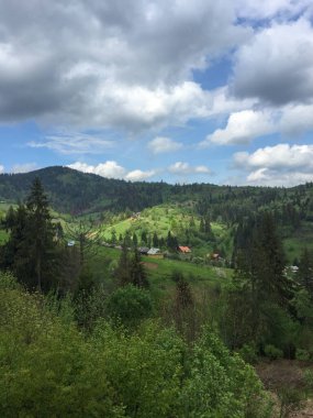 Bereketli yeşil tepeler parçalı bulutlu bir gökyüzünün altında arazi boyunca uzanıyor. Vadide ağaçlar ve dağ manzarasıyla çevrili birkaç ev görülebilir..