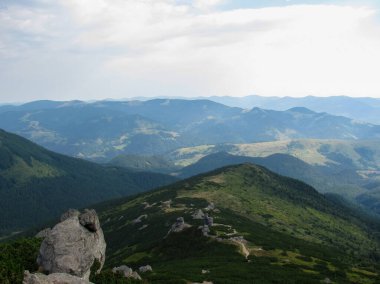 Parçalı bulutlu bir gökyüzünün altında yuvarlanan tepeleri ve uzak dağları gösteren nefes kesici bir manzara. Yeşil bitki örtüsü, bu huzurlu yerdeki doğanın güzelliğini vurguluyor..