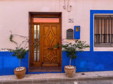 Bu davetiye girişi, canlı mavi bir duvara karşı güzel yapılmış ahşap bir kapı içeriyor. İki saksı bitkisi biraz yeşillik katarak, hoş geldin atmosferini güçlendirir..