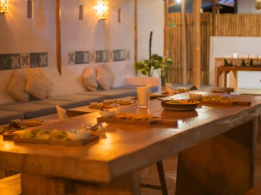 Yemek için ahşap masa seti olan samimi bir restoranın seçici odak noktası. Duvarlardaki sıcak ışık ve çerçeveli sanat eserleri davetkar bir atmosfer yaratıyor. Bakış açısı odanın derinliğini ve düzenini gösteriyor..