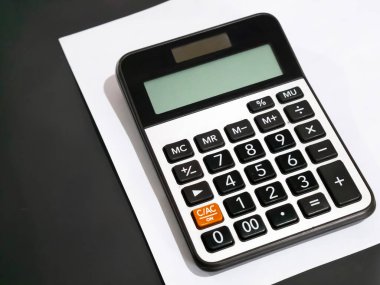 Beyaz bir kağıt üzerinde bir hesap makinesinin yakın çekimi. Görünür anahtarlar arasında sayısal anahtarlar (0-9), temel işlem anahtarları (toplama, çıkarma, çarpma, bölme) ve özel fonksiyon tuşları bulunmaktadır..