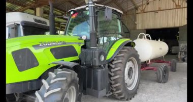 Yeşil ve beyaz traktör bir tarlada park halinde. Traktör beyaz bir tankın yanında.
