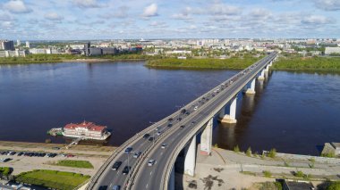 Köprü, arka planda bir şehir bulunan bir nehri kapsar. Köprü arabalarla dolu ve bir tekne nehre yanaştı.