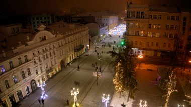 Şehir caddesi, ortasında büyük bir Noel ağacı olan bir gece. Sokaklar ışıklarla aydınlanıyor ve insanlar etrafta dolanıyor.