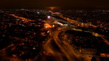Geceleri arka planda büyük bir bina olan bir şehir. Şehir ışıklarla aydınlanıyor ve gökyüzü bulutlu.