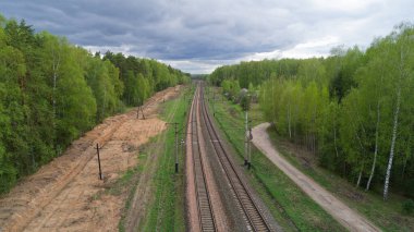 Arka planda orman olan tren rayları. Tren hareket halinde değil. Gökyüzü bulutlu.