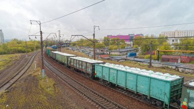 Tren raylarda ilerliyor, arkasında da birkaç araba var. Tren arka planında yüksek binalar olan bir şehirle çevrilidir. Sahne biraz kasvetli.
