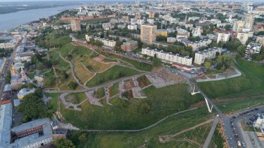 Şehir manzarası yukarıdan, ortada büyük yeşil bir tepe var. Tepe binalar ve bir nehirle çevrilidir. Şehir hareketlilikle dolup taşıyor, etrafta bir sürü insan dolaşıyor. Nizhny Novgorod