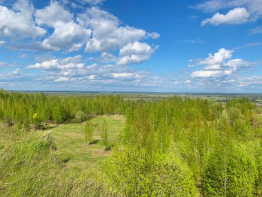 Ağaçları ve açık mavi gökyüzü olan yemyeşil bir arazi. Gökyüzü bulutlarla kaplı, ortama huzurlu ve huzurlu bir atmosfer veriyor.