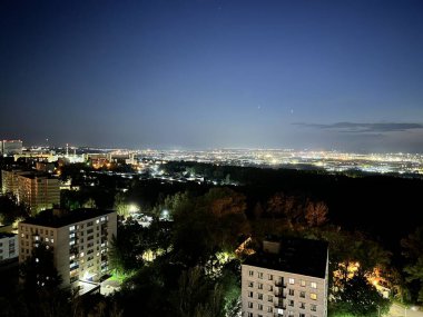 Geceleri gökyüzü karanlık ve bulutlu bir şehir silueti. Şehir, binalardan ve sokak lambalarından gelen ışıklarla aydınlanıyor.