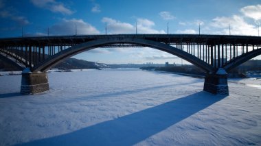 Köprü, yerde karla kaplı bir nehri kaplar. Köprü büyüktür ve nehrin tüm genişliğini kapsar.