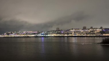 Arka planda bir şehir olan karanlık bir gece gökyüzü. Su sakin ve şehrin ışıkları suyu yansıtıyor.