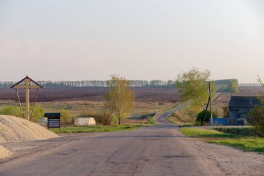 Üzerinde tabela olan toprak bir yol. Uzakta bir ev var. Yol boş.