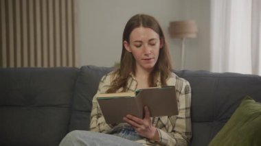 Dairesinde kitap okuyan bir kadın, bir kitabın sayfalarını çeviriyor.