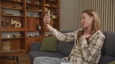 Video konferansında konuşan genç kadın Smartphone 'dan arıyor, Apartmandaki koltukta oturuyor.