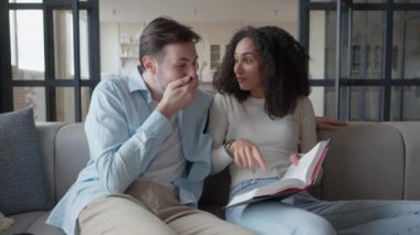 Genç bir çift oturma odasında kağıt kitap okuyor ve gülümsüyor, sevimli bir aile, evdeki kanepede birlikte vakit geçiriyorlar, yüksek kaliteli 4K görüntüler.