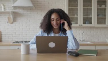 Genç kıvırcık Afrikalı kadın telefonuyla içeride dizüstü bilgisayarın önünde konuşuyor, yüksek kalite 4K görüntü.