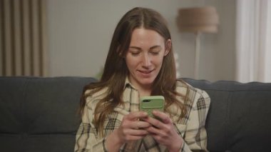 Smartphone 'da gülümseyen kadın evde dinlenirken online sohbetin tadını çıkarıyor. Mutlu Mesajlaşma, Sosyal Medya, Dijital İletişim, Konforlu Atmosfer, Günlük Yaşam Tarama, İnternet Taraması