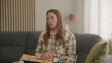 Üzgün kadın pizzayla koltukta oturuyor, düşünceli ve düşüncelerde kaybolmuş görünüyor. Duygusal Anlar, Derin Düşünme, Rahatlatıcı Yemek, Düşünceli İfade, Ev Atmosferi, Rahatlamış Ayar, Günlük Yaşam Tarzı