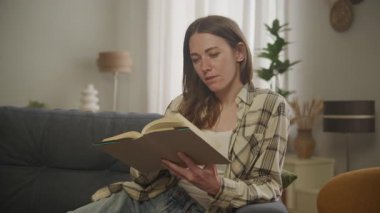 Evde rahat rahat otururken kitap okuyan ve sayfaları çeviren genç bir kadın. Rahat atmosfer, boş zaman, eğlenen edebiyat, günlük yaşam tarzı huzurlu an, dinlendirici ev, modern iç mekan.