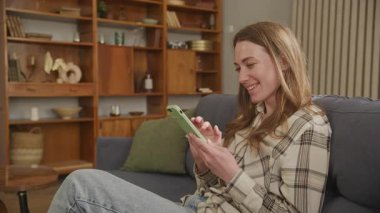 Koltukta oturan gülümseyen kadın, evde akıllı telefondan mutlu mesajlar okuyor. Dijital İletişim, Sosyal Medya Sohbeti, Pozitif Duygu, Konforlu Atmosfer, Modern Teknoloji, Rahat Yaşam Tarzı