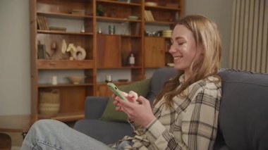 Mutlu kadın kanepeye yaslanıyor, neşeyle gülüyor ve rahatlatıcı anın tadını çıkarıyor. Pozitif Duygular, Konforlu Atmosfer, Neşeli İfade, Sıradan Yaşam Tarzı, Ev Rahatlığı, Yüksek Kaliteli 4K Görüntü