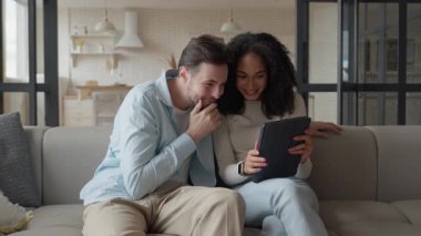 Tablet okuyup iyi haberler alırken ırklar arası bir çift gülüyor. Beyaz adam ve Afro-Amerikan kadın sıcak bir evde olumlu gelişmelerden keyif alıyorlar. Mutluluk kavramları için mükemmel