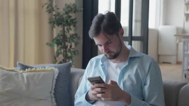 Kafası karışan adam, akıllı telefondan koltukta otururken beklenmedik bir mesaj okuyor. Şaşırmış İfade, Kalkmış Kaşlar, Odaklanmış Bakış. Sıradan Atmosfer, Beklenmedik Haberler. Yüksek kalite 4k görüntü