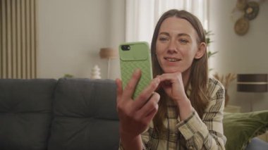 Green Case 'le görüntülü telefon görüşmesi yapan Özel Bir Kadın. Konuşurken gülümsemek ve doğal tepkiler vermek, sıcak ışıklarla sıcak evde oturmak, sıradan kıyafetler giymek.