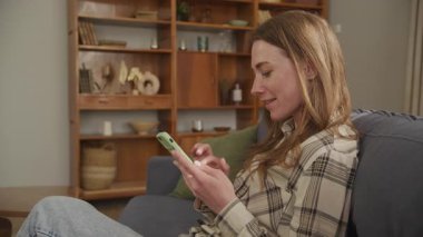 Genç bir kadın rahat bir şekilde koltukta oturuyor, akıllı telefondan mesaj yazmakla meşgul. Yüksek kalite 4k görüntü