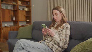 Neşeli kadın telefonu kullanıyor, gülümsüyor ve rahat koltukta dinleniyor. Mutlu Genç Kadın Evde Gündelik Anın keyfini çıkarıyor, Çevrimiçi Sohbete daldı. Rahat atmosfer, pozitif titreşimler, Modern Teknoloji