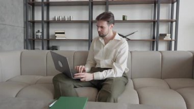 Dizüstü bilgisayarda çalışan ciddi bir adam kucağında rahat bir şekilde koltukta oturuyor. Uzaktan Çalışma, Üretim, Konsantrasyon, Modern İçişleri, Dijital Göçebe, İş Dünyası. Yüksek kalite 4k görüntü