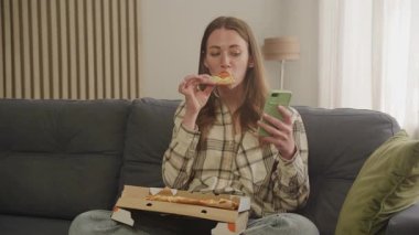 Pizzayı seven ve evde rahat bir şekilde otururken akıllı telefon kullanan bir kadın. Rastgele Akşam Rahatlama Sahnesi, Lezzetli Fast Food, Modern Yaşam Tarzı, Dijital Boş Zaman, Gerçek Anlar, Konforlu Ev Keyfi