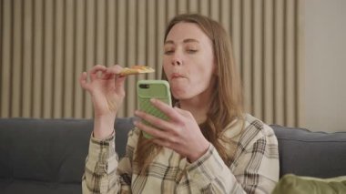 Genç bir kadın koltukta dinlenirken pizza yiyor ve akıllı telefonlara bakıyor. Modern kadının evde boş vakitlerinde konfor yemeği ve dijital eğlencenin keyfini çıkardığı günlük yaşam tarzı sahnesi.