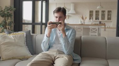 Parlak Modern Oturma Odası İçi 'nde Mobil Oyun Oynayan Adam. Rahat adam Smartphone Oyun Odaklandı, Evde Rahatça Oturma, Konsantrasyon Gösterme, Yüksek Kaliteli 4K görüntü