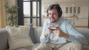 Çevrimiçi oyun oynayan, kulaklık takan, konuşan ve rakiplerine karşı kazanan bir adam. Oyuncu, Konforlu Modern Ev Oturma Odası 'ndan Kendine güven ve Neşe İfade Ediyor.