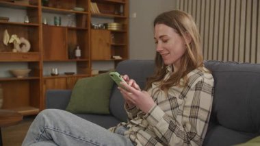 Smartphone 'da parıldayan kadın evde rahat rahat otururken mesaj atıyor. Huzurlu Ev Atmosferi, Doğal Işık, Rahat Kıyafet, Günlük Teknoloji Kullanımı ve Ev Kullanımı Konsepti